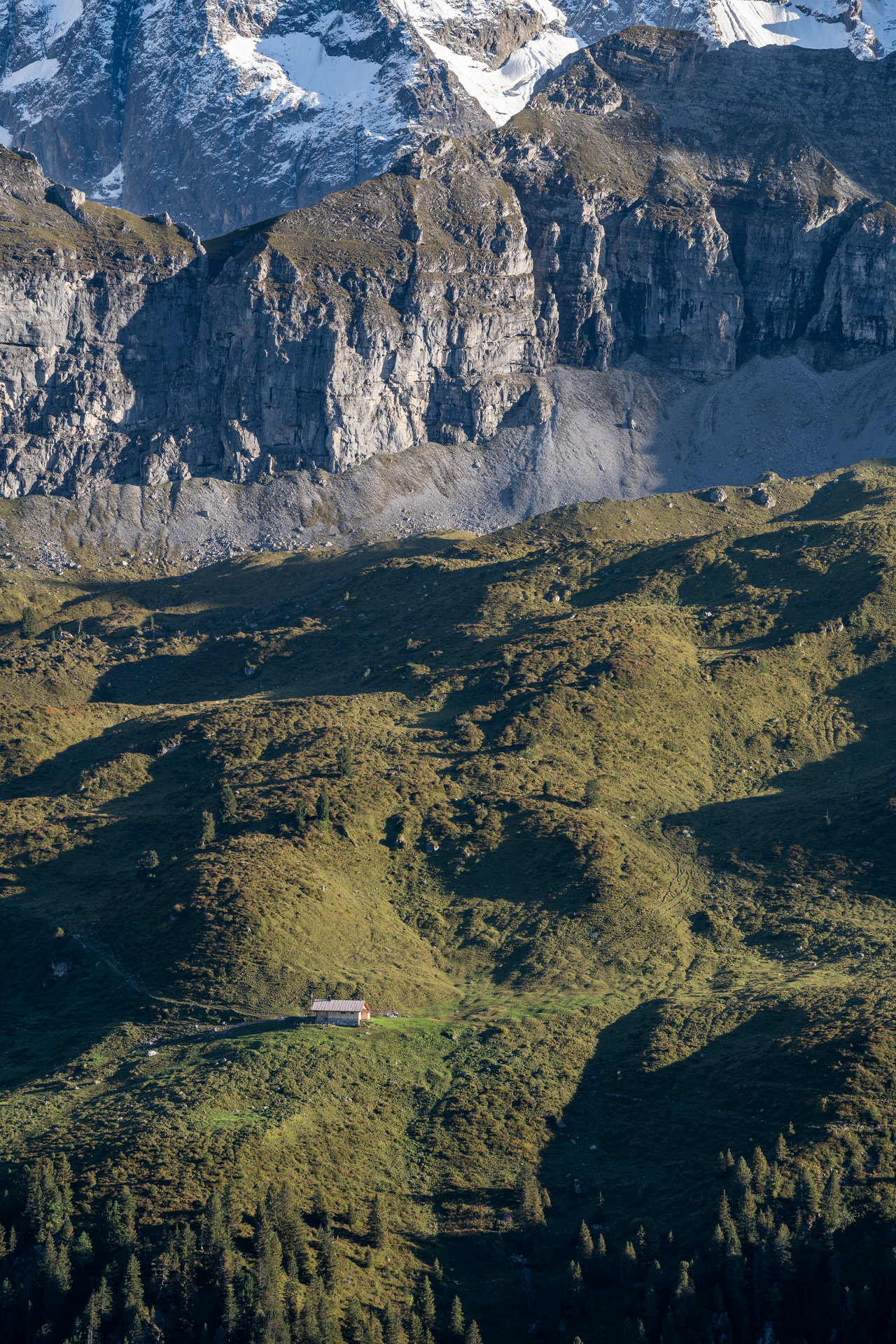 A single tiny house in the bottom left corner surrounded by huge hills and mountains. It dwarfs in comparison to a surrounding landscape.