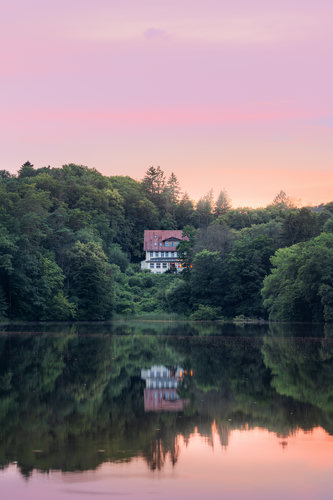 Old small hotel stands on the shore of the lake. It is surrounded by thick woodland. Above, there is a beatifyl firely-red sunset sky. Sky, hotel and the trees are reflecting in the lake.